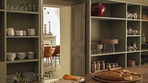 A kitchen with open shelving displaying dishes and jars, a wooden table with bread and plates, and a view into a dining area with chairs. - Dimora Santagatha  (Noto)