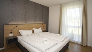 Ein modernes Hotelzimmer mit einem großen Bett, einem Holz-Kopfteil, einem Nachttisch und einem großen Fenster mit transparenten Vorhängen.