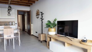 A modern kitchen with white cabinets, a wooden dining table, and chairs. There is a large flat-screen TV on a wooden stand, a potted plant, and a wall-mounted clock. - Ca' Linda By Case al Lago (Desenzano del Garda)