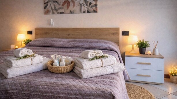A neatly made bed with a purple quilt, two side tables with lamps, a small plant, and a basket of toiletries. - Ca' Linda By Case al Lago (Desenzano del Garda)