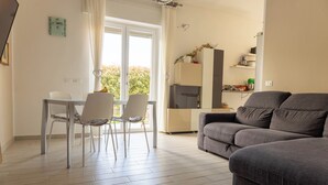 En moderne stue med en grå sofa, et spisebord med stole og et køkken med køleskab og hylder.