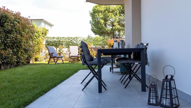 En udendørs patio med et bord og stole, omgivet af en hæk og en bygning i baggrunden.