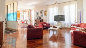 A spacious living room with wooden flooring, red sofas, a coffee table, a TV, and a dining area with chairs and a table.