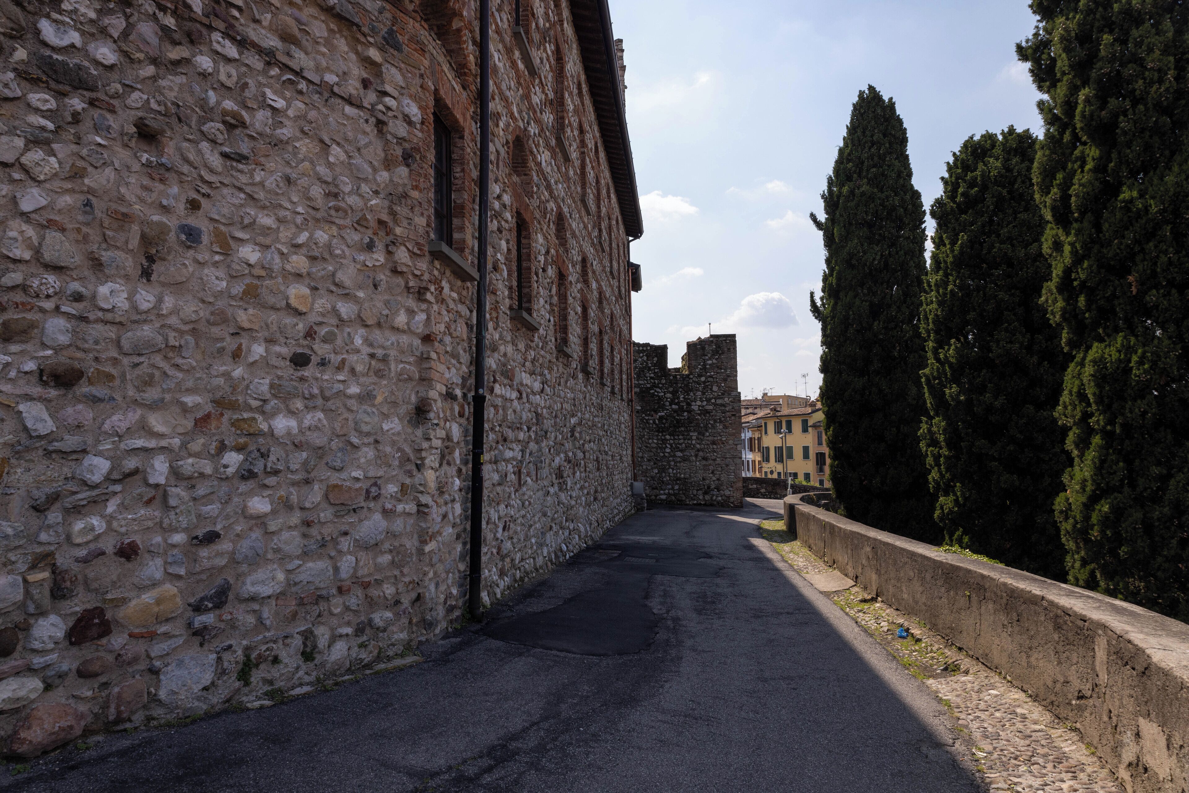 A narrow street with stone walls on one side and a row of trees on the other.