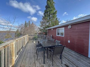 Une terrasse en bois avec une table et des chaises, un bâtiment rouge et une vue sur des arbres et une étendue d’eau.