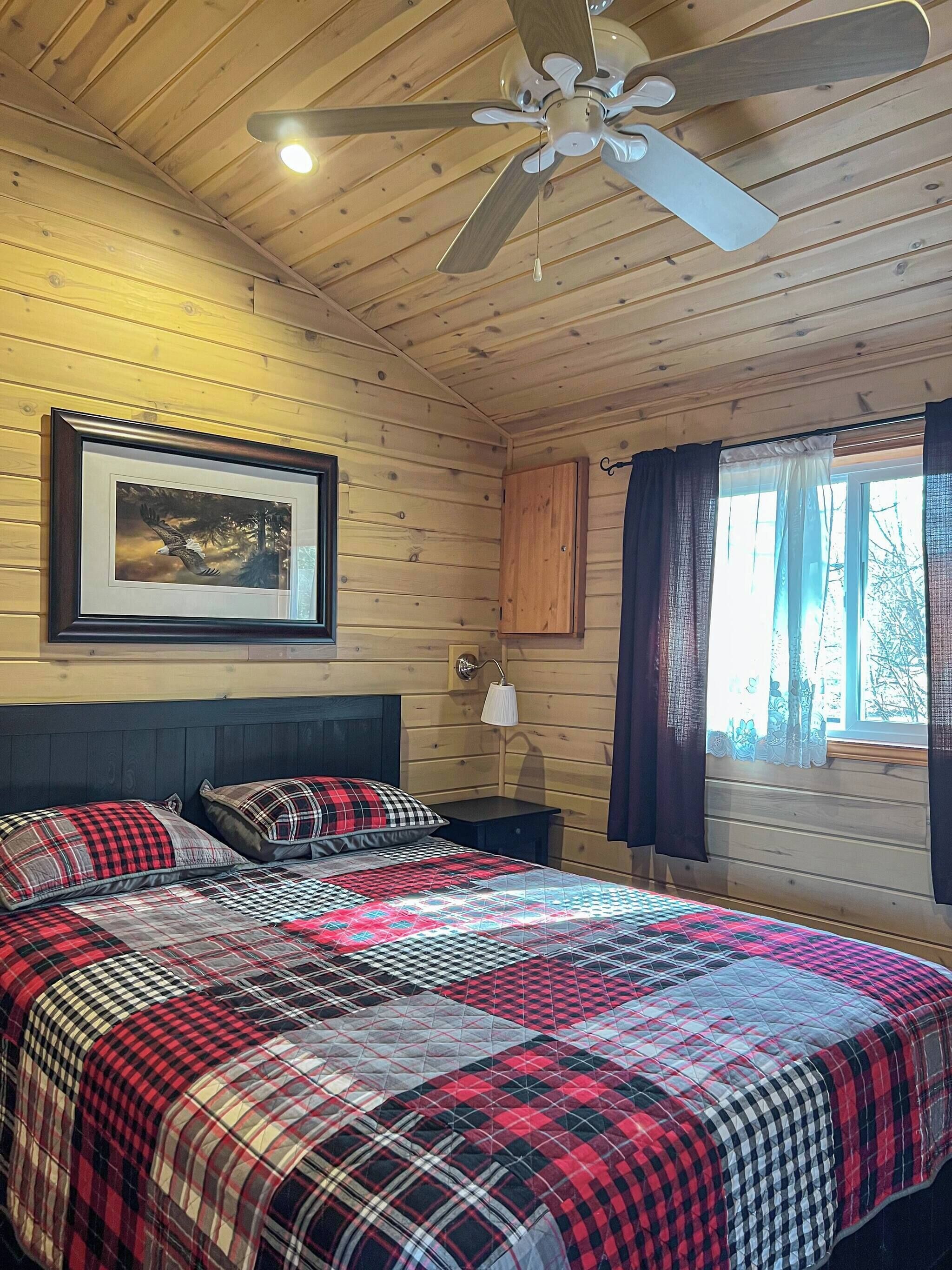 A wooden cabin bedroom with a bed, a ceiling fan, a framed picture, a nightstand, and a window with curtains.