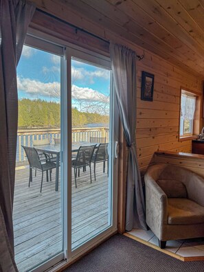 A wooden cabin interior with a sliding glass door leading to an outdoor deck with chairs and a table.