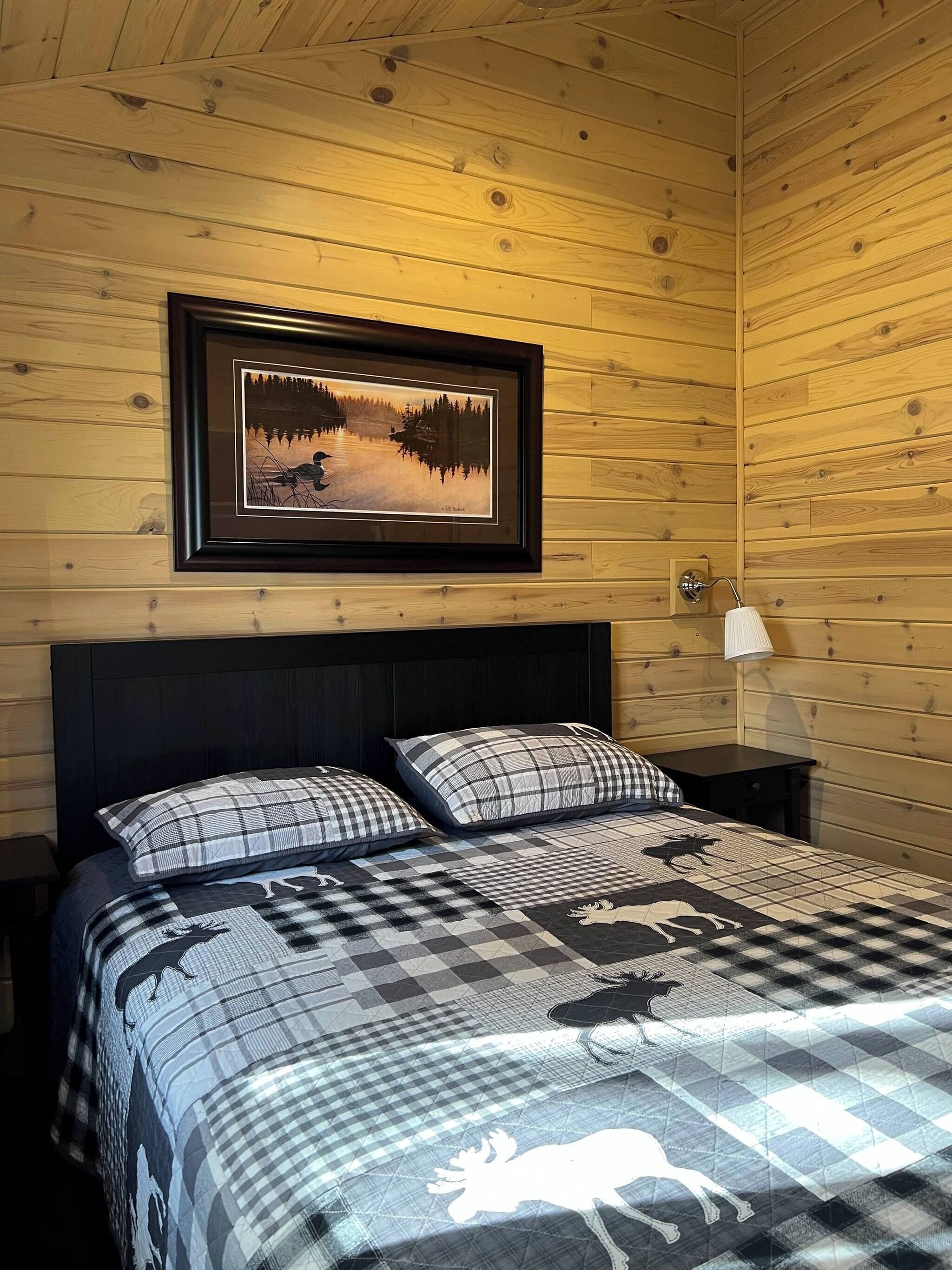 A wooden cabin bedroom with a bed, two pillows, a framed picture, a nightstand, and a lamp.