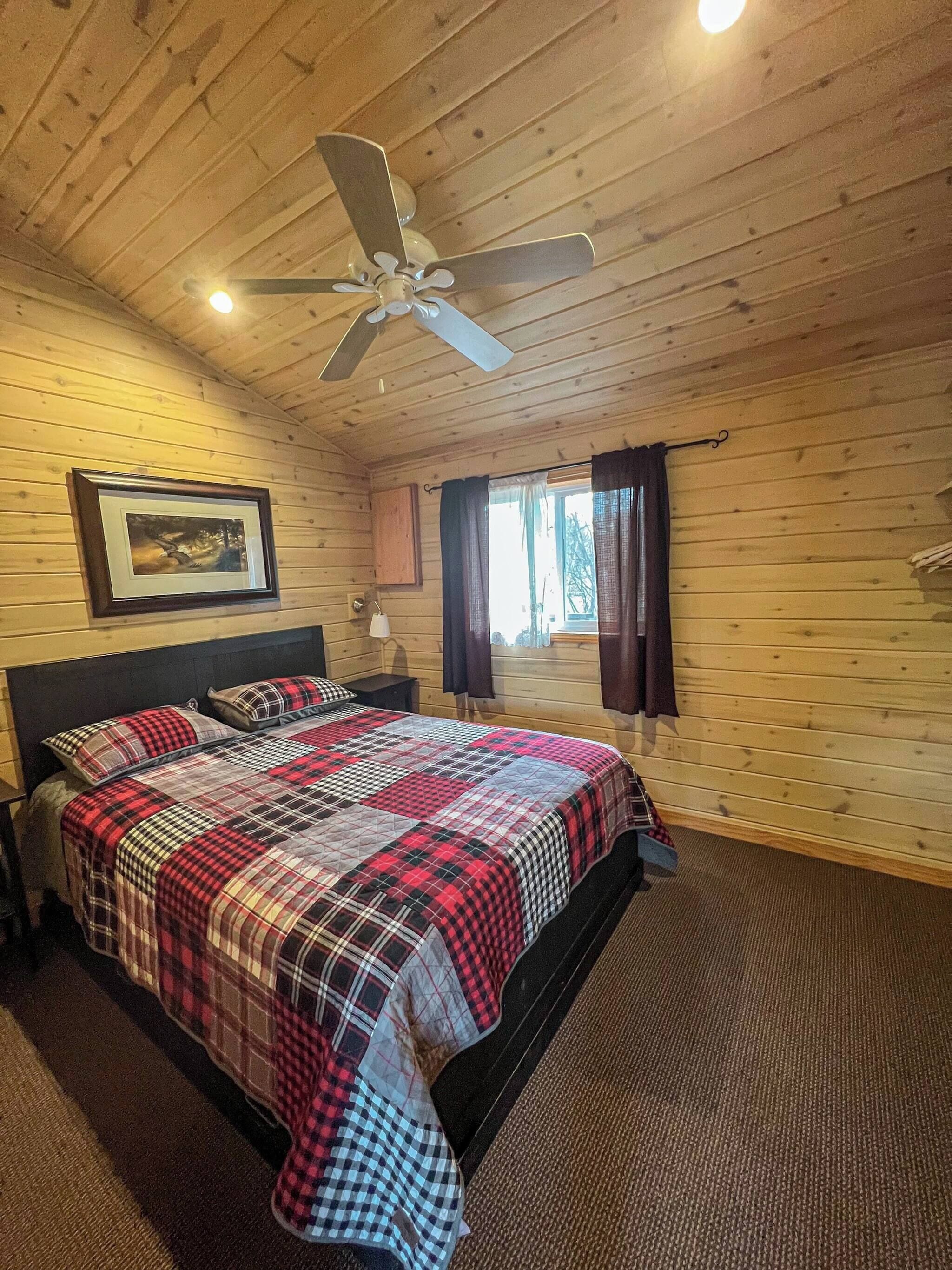 A wooden-paneled bedroom with a bed, a ceiling fan, a framed picture on the wall, and a window with curtains.