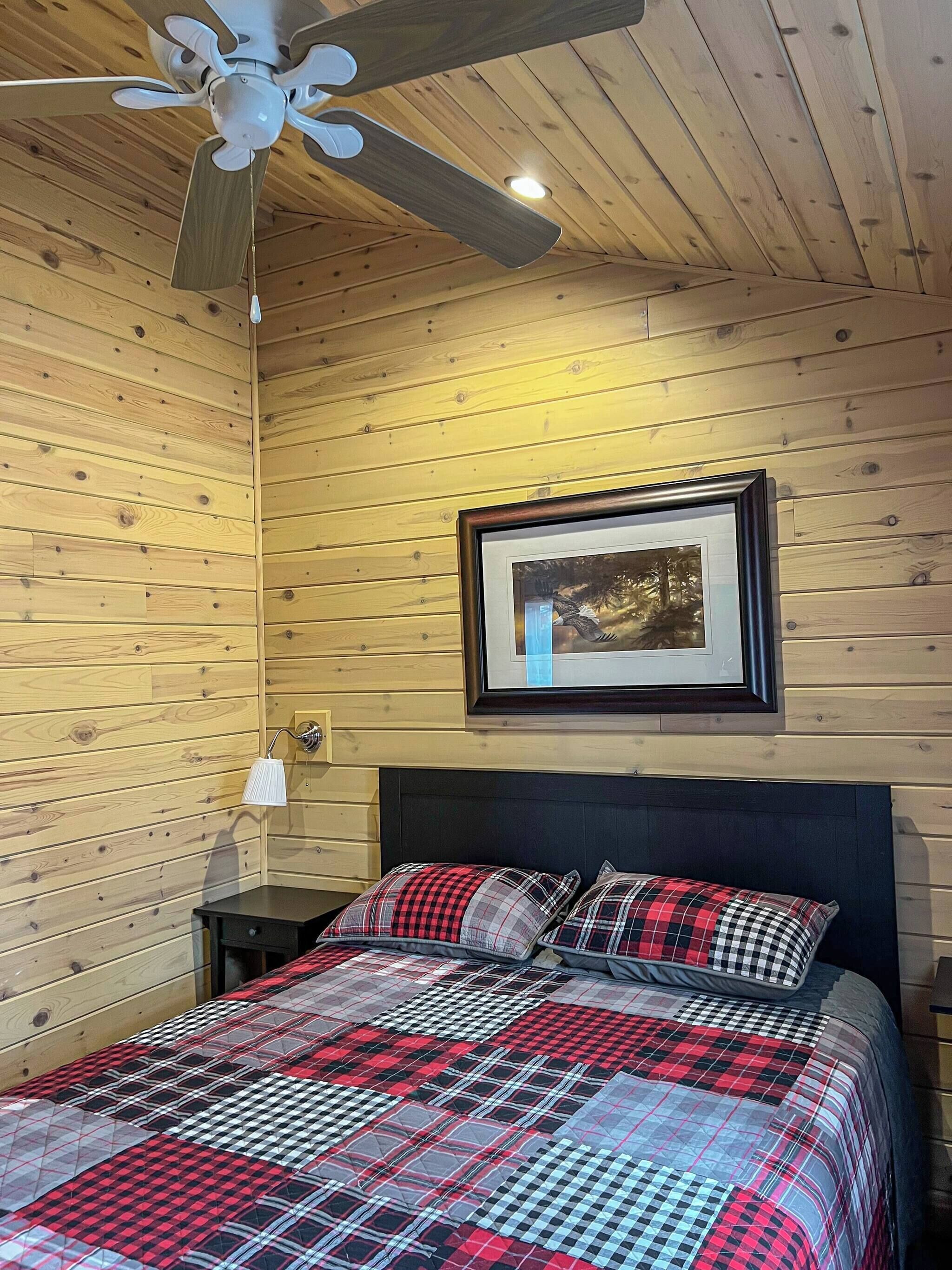 A wooden-paneled room with a bed, a ceiling fan, a framed picture on the wall, and a bedside lamp.
