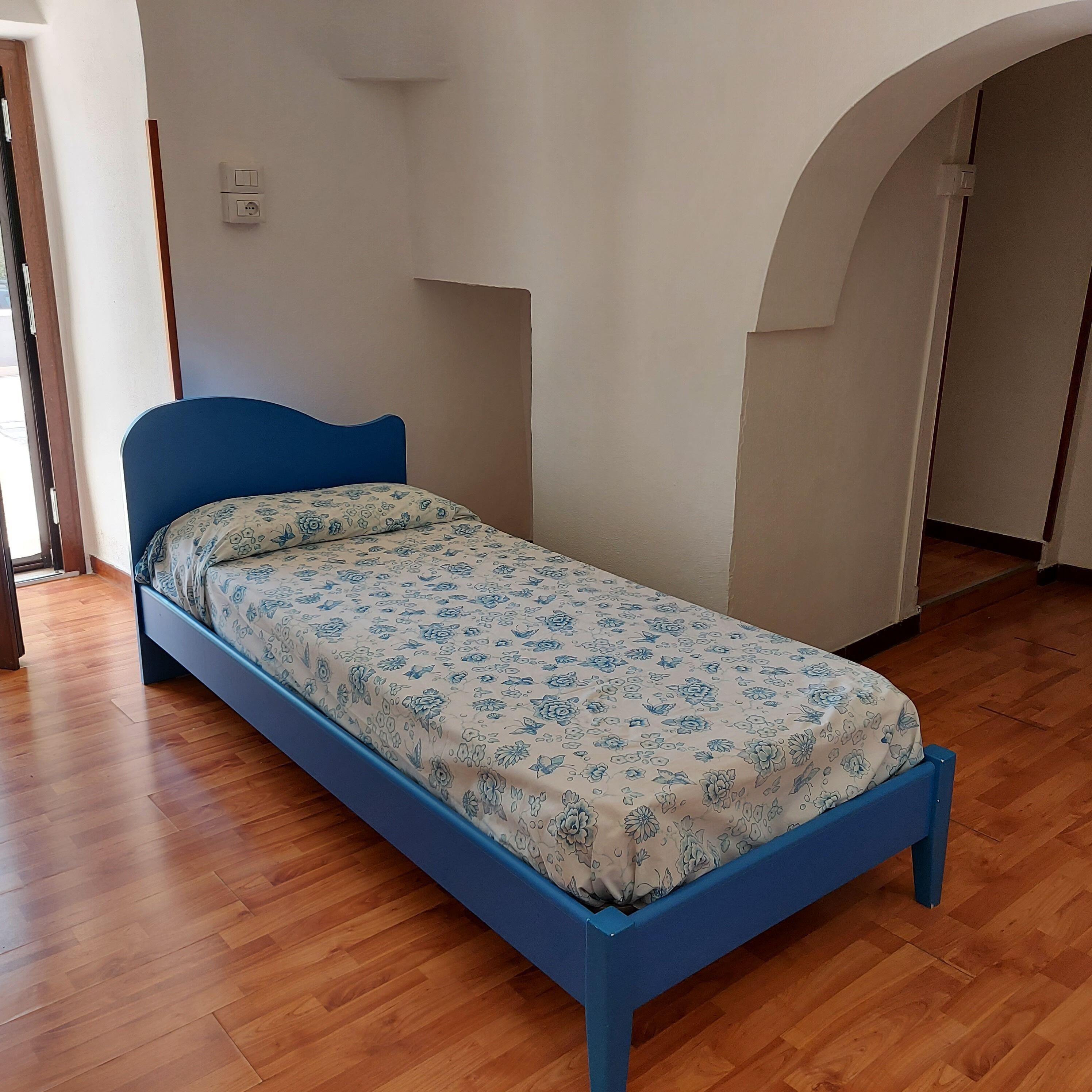 Una cama individual con una colcha estampada en un cuarto con piso de madera y paredes blancas.