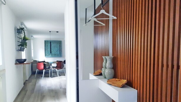 Un interior moderno con un panel de madera, una estantería blanca con objetos decorativos, y un comedor con mesa y sillas.