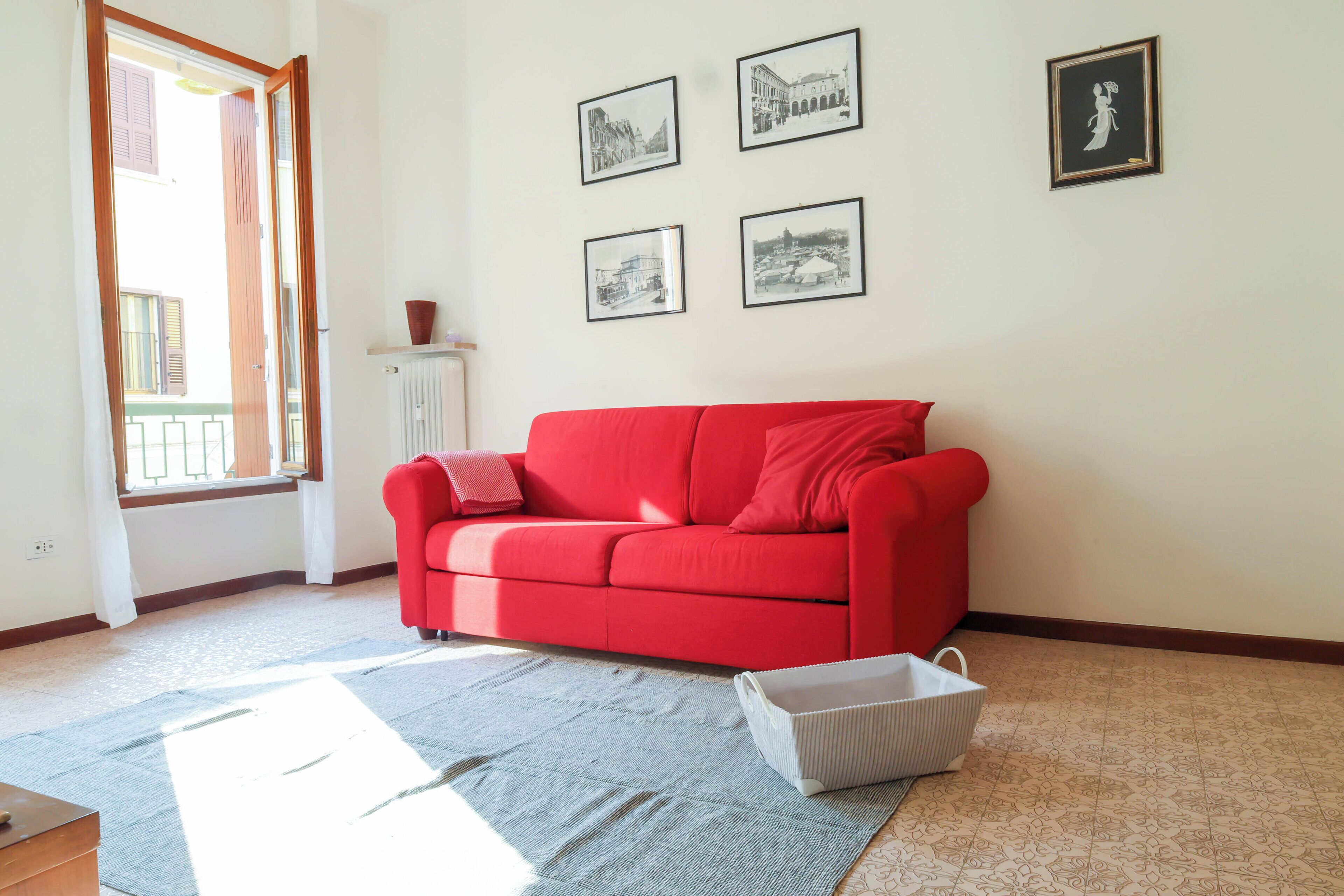 A red sofa in a room with a window, a radiator, and four framed pictures on the wall.