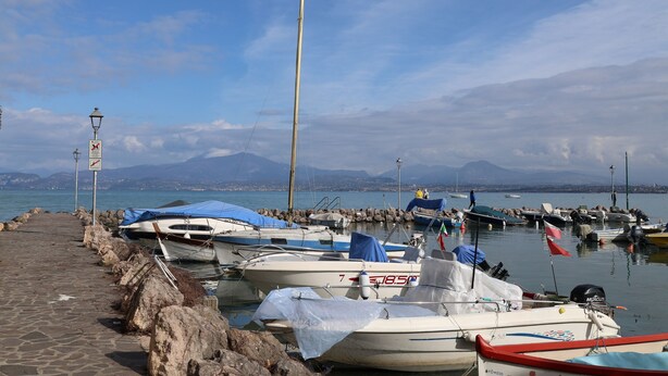 Un port de plaisance avec des bateaux amarrés le long d’une promenade dallée, un ciel dégagé et des montagnes en arrière-plan.