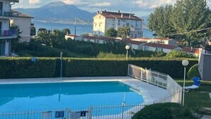 Une piscine entourée d’une clôture blanche, bordée de verdure et offrant une vue dégagée sur la mer et les montagnes en arrière-plan.