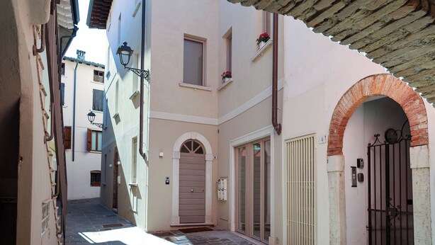 A narrow alleyway with a building entrance featuring a wooden door, a small window with flowers, and a metal gate. - Vicolo dei Signori By Case al Lago (Desenzano del Garda)