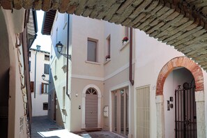 Une ruelle étroite, avec l’entrée d’un immeuble caractérisée par une porte en bois, une petite fenêtre ornée de fleurs et un portail en métal.
