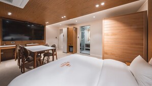 A modern hotel room with a wooden ceiling, a large flat-screen TV, a dining area with a table and chairs, and a large bed with a logo on the mattress. - Gridt Hotel (Gyeongju)