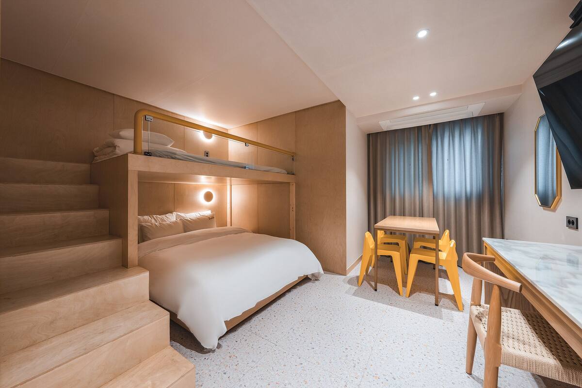 A modern hotel room with a bunk bed, a desk with chairs, and a staircase.