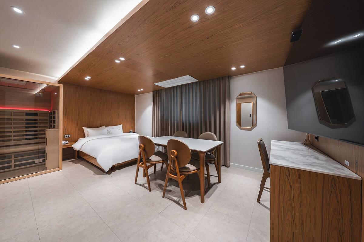 A modern bedroom with a wooden ceiling, a bed with white linens, a dining table with chairs, and a small desk area.