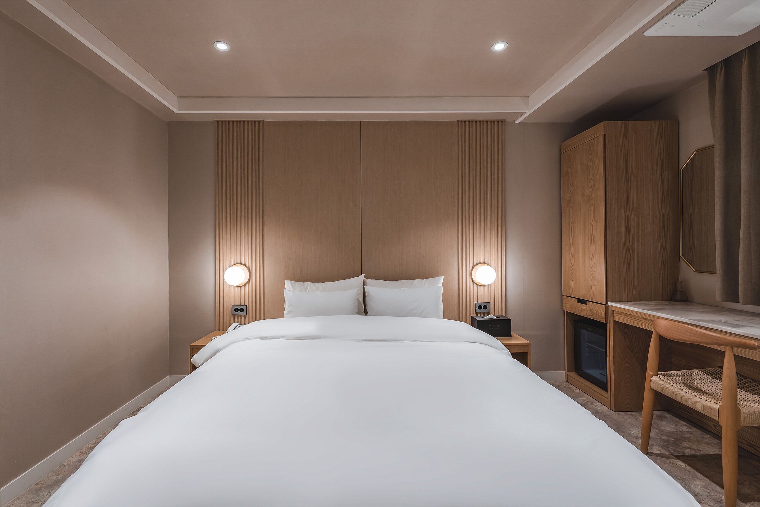 Una habitación de hotel moderna con una cama grande, cabecera de madera, mesitas de noche con lámparas y un pequeño escritorio de madera con una silla.