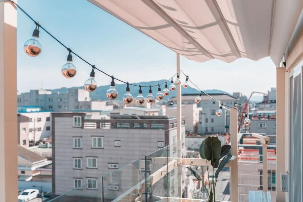 Un balcon orné d’une guirlande de petites lampes sphériques, doté d’une balustrade en verre et offrant une vue sur les bâtiments de la ville.
