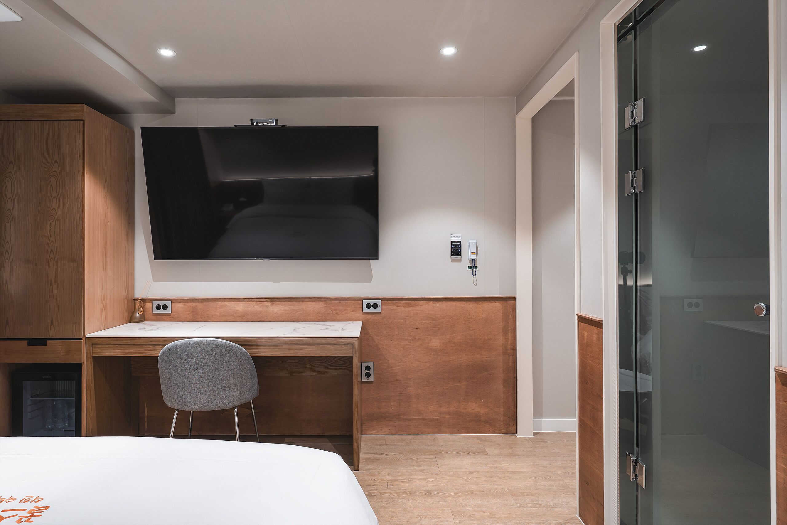 A modern hotel room with a bed, a desk, a chair, a television, and a glass door.