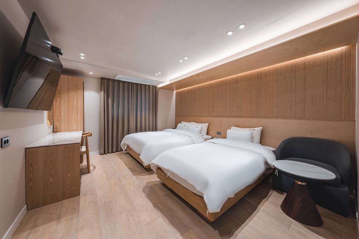 A modern hotel room with two beds, a small desk, a chair, and a TV mounted on the wall.