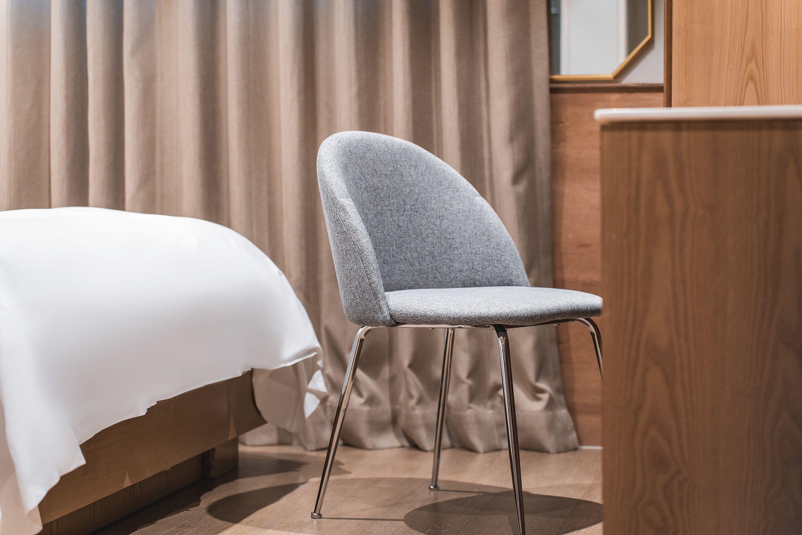 Un lit bien fait, recouvert d’une couette blanche, une chaise rembourrée de couleur grise avec des pieds en métal et un bureau en bois, le tout dans une pièce dont le fond est constitué d’un rideau.