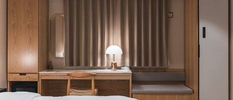 A compact hotel room with a bed, a desk with a chair, a lamp, a wardrobe, and a built-in refrigerator.