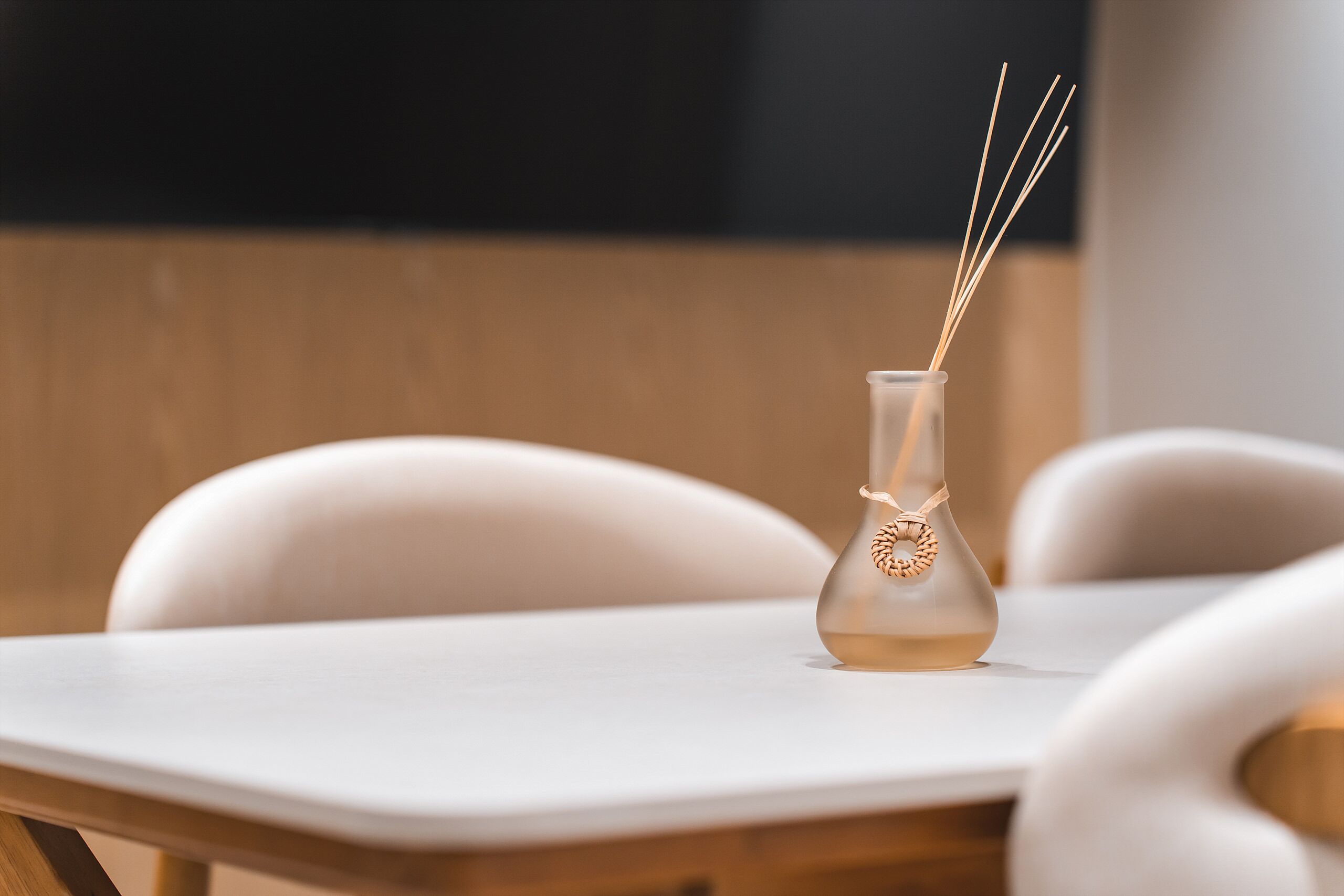 A white table with a clear glass bottle containing reed sticks, set on a wooden chair.