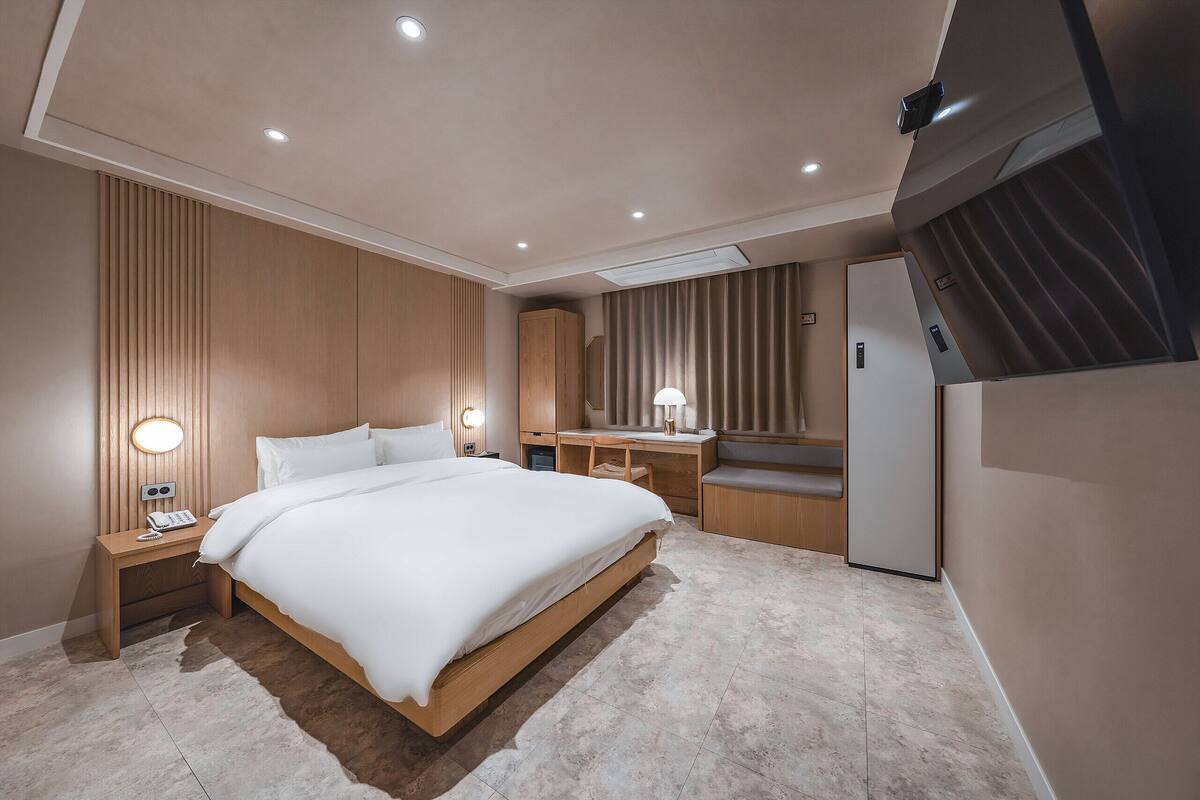 A modern hotel room with a large bed, a desk, a chair, a wardrobe, and a mirror.