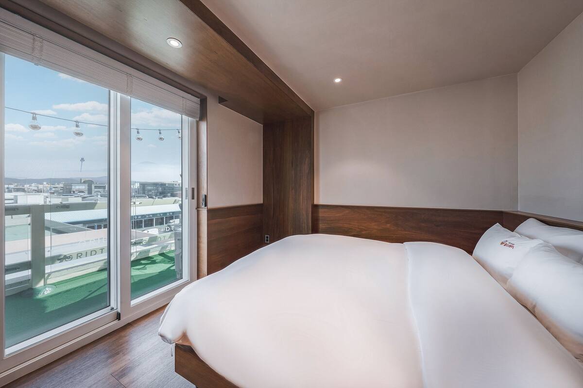 A modern hotel room with a large bed, wooden headboard, and a sliding glass door leading to a balcony with a city view.