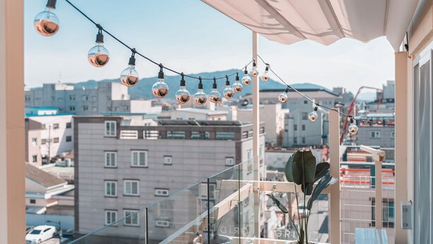 A balcony with a string of hanging lights, a potted plant, and a view of city buildings. - Gridt Hotel (Gyeongju)