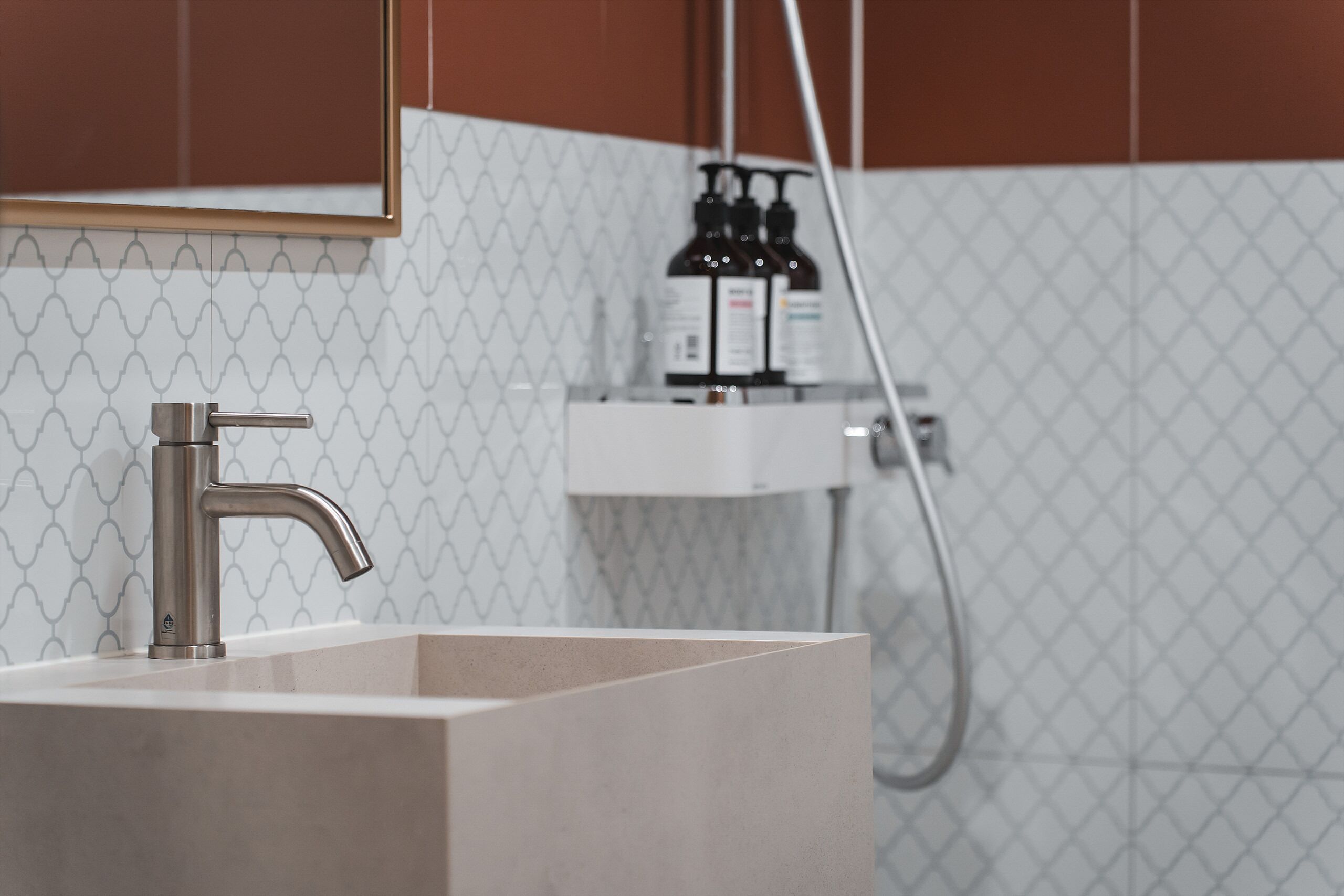 A bathroom with a modern sink, a faucet, and a mirror. There is a showerhead and a bottle of shampoo on the wall.