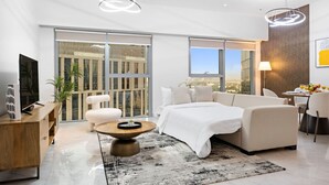 A modern living room with a sofa, a coffee table, and a TV. There is a large window with a city view.