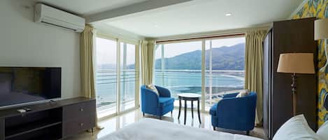 A hotel room with a large bed, a TV, a desk, two chairs, a lamp, and a view of the sea.