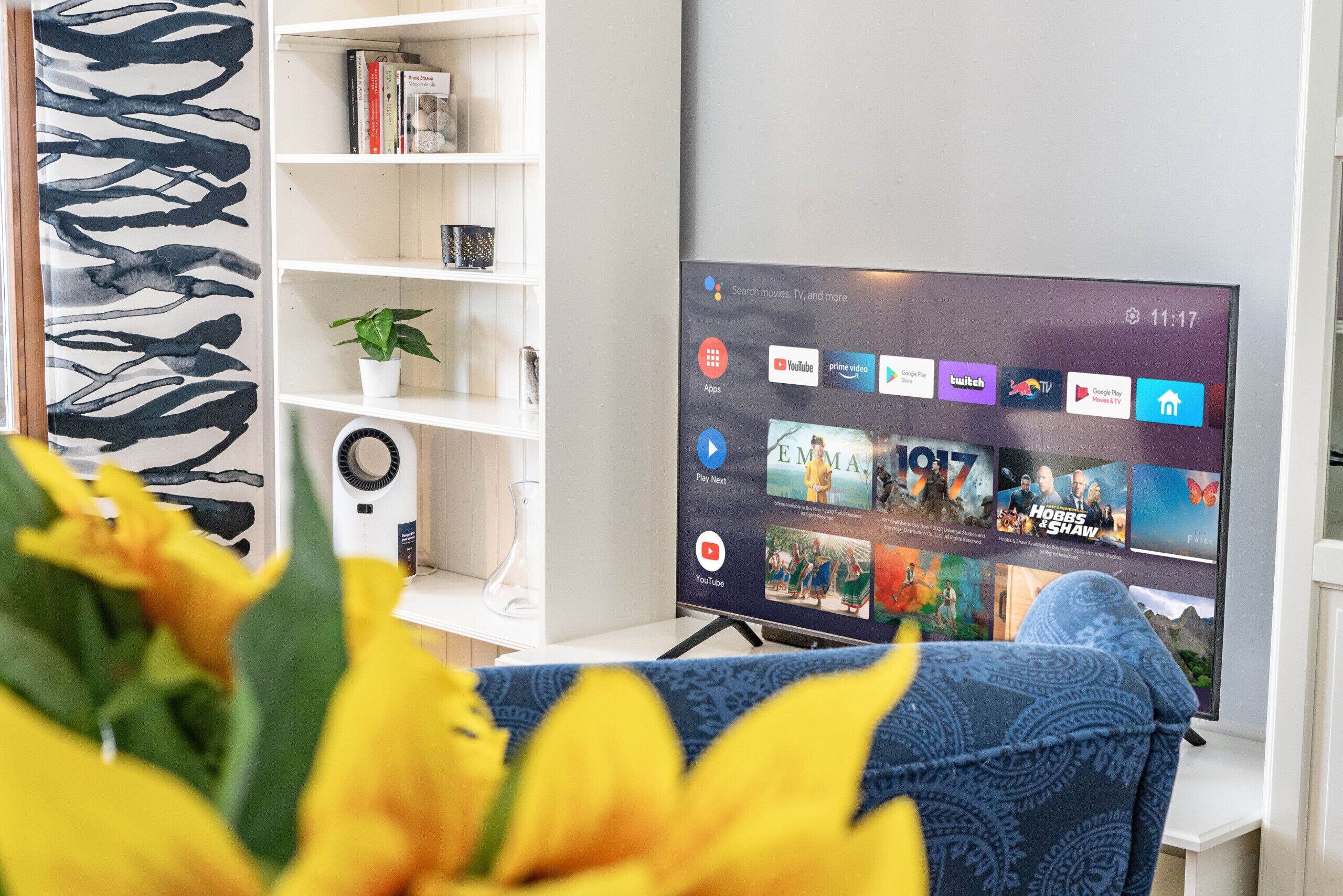 Olohuoneessa on litteänäyttöinen televisio, jossa näkyy suoratoistopalvelun käyttöliittymä, valkoinen kirjahylly, jossa on kirjoja ja kasvi, sekä sinikuvioinen sohva.
