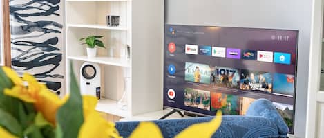 Olohuoneessa on litteänäyttöinen televisio, jossa näkyy suoratoistopalvelun käyttöliittymä, valkoinen kirjahylly, jossa on kirjoja ja kasvi, sekä sinikuvioinen sohva.