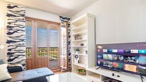 A modern living room with a glass coffee table, a black sofa, and a large flat-screen TV mounted on the wall. There is a sliding glass door leading to a balcony with a view of the outdoors.