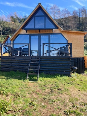 Una cabaña de troncos con un ventanal grande, una terraza de madera y una parrilla.
