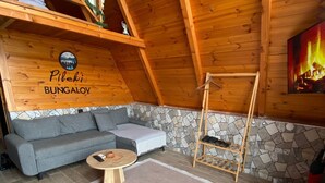 A cozy room with a stone wall, a gray sofa, a wooden table, and a fireplace. - Pileki Bungalov (Iyidere)