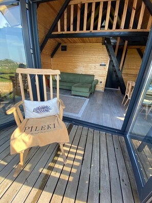Una terraza de madera con una silla y un sofá dentro de una cabaña de madera con una pared de vidrio.