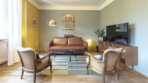 Un salon avec un canapé en cuir marron, deux fauteuils en osier, une table basse en verre, un téléviseur posé sur un meuble en bois et une lampe sur pied.