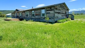 Une cabane en bois rustique, dotée d’un porche et d’un escalier, entourée d’un champ verdoyant et avec des montagnes en arrière-plan.