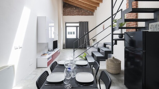 A modern kitchen with a black refrigerator, a dining area with a table set for four, and a staircase with a brick wall.
