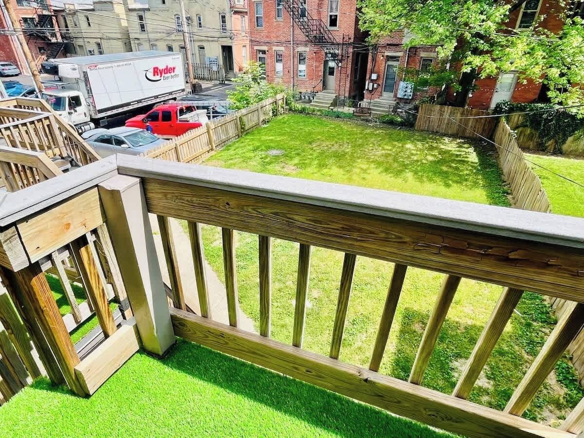 Un jardin à l’arrière d’une maison, avec une terrasse en bois, de l’herbe artificielle verte et un bâtiment en briques en arrière-plan.