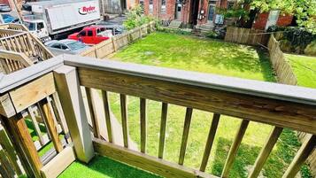 Un jardin à l’arrière d’une maison, avec une terrasse en bois, de l’herbe artificielle verte et un bâtiment en briques en arrière-plan.