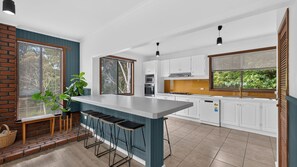 A modern kitchen with a central island, bar stools, and a view of greenery outside. - Treetop Retreat House | Spacious Family Getaway (Metung)