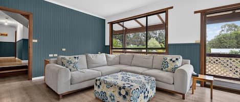 Un salon moderne avec un canapé d’angle, un pouf à motifs et une grande fenêtre donnant sur un paysage verdoyant.